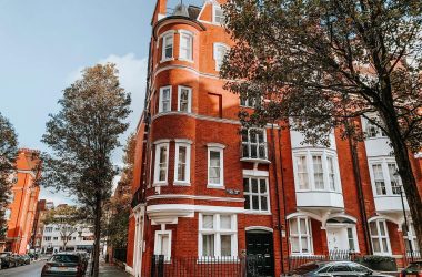 A red brick building with multiple windows and a rounded corner, surrounded by trees in London.