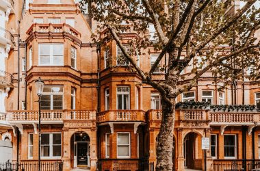 Row of ornate Victorian-style terraced townhouses with bay windows and balconies, partially shaded by a large tree on a residential street.