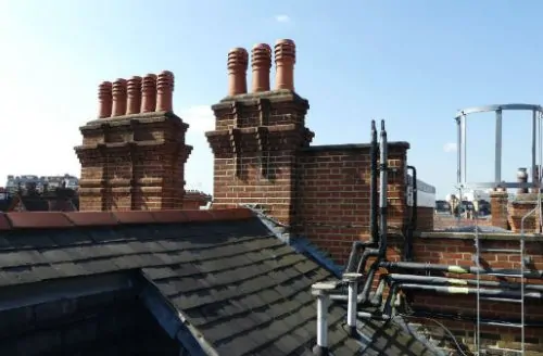 Rooftop view with slate tiles, brick chimneys, chimney pots, and metal pipework against a clear sky.