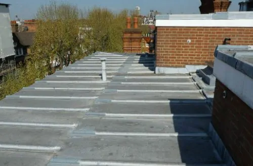 Flat rooftop with grey waterproofing, low parapet walls, a brick chimney, and trees visible in the background.
