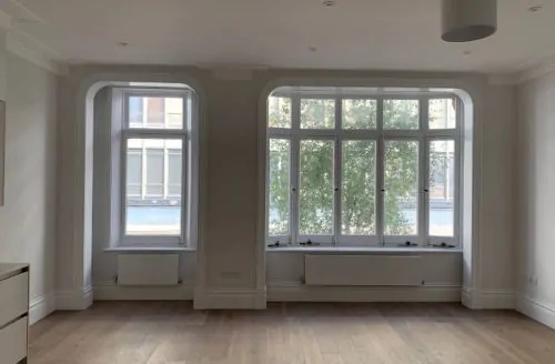 Bright, empty room with large windows allowing natural light, showcasing clean walls and hardwood floors .