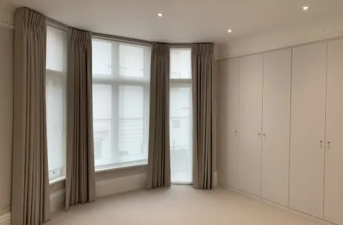 Empty bedroom with light carpet, neutral walls, tall windows with curtains, fitted wardrobes, and recessed ceiling lights.