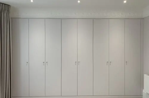 Empty room with fitted white wardrobes, light flooring, plain walls, and recessed ceiling lights.
