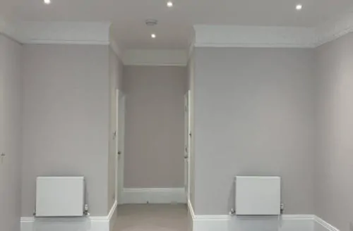 Empty neutral room with light flooring, recessed ceiling lights, grey walls, two radiators, and a small alcove or doorway at the back.