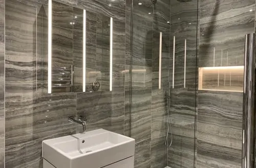 Modern bathroom with grey marble-effect tiles, a white wall-mounted sink, large mirror, and vertical light strips.
