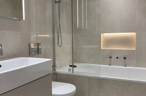 Modern bathroom with grey wall tiles, a bathtub with shower fittings, a white sink, toilet, and illuminated mirror cabinet.