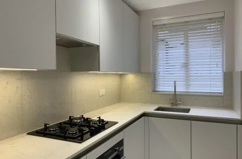 Compact modern kitchen with grey cabinets, light countertops, built-in oven, gas hob, and a window with white blinds.