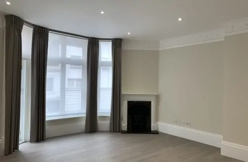 Empty living room with light wood flooring, a bay window with dark curtains, neutral walls, recessed ceiling lights, and a small fireplace.