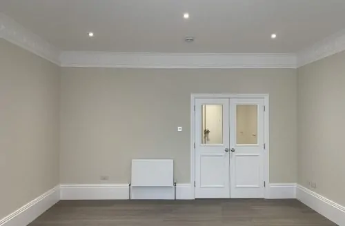 Empty room with grey walls, light flooring, recessed ceiling lights, a radiator, and white double doors with glass panels.