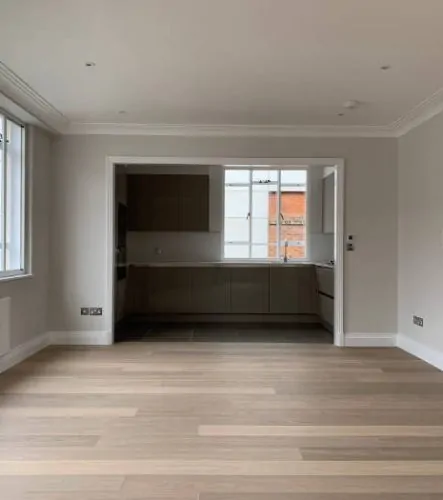 Empty living space with light wood flooring, leading into a modern kitchen, part of a recent building refurbishment project.