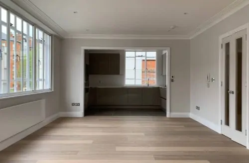 Empty living space with light wood flooring, leading into a modern kitchen, part of a recent building refurbishment project.