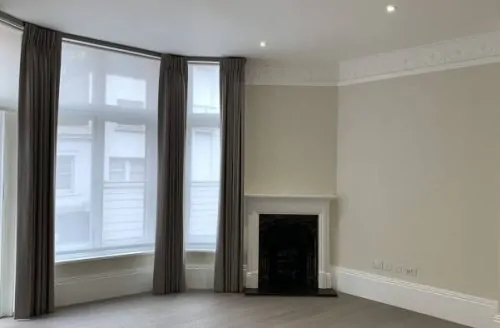 Empty room with a bay window, dark curtains, neutral walls, light flooring, and a small black fireplace.