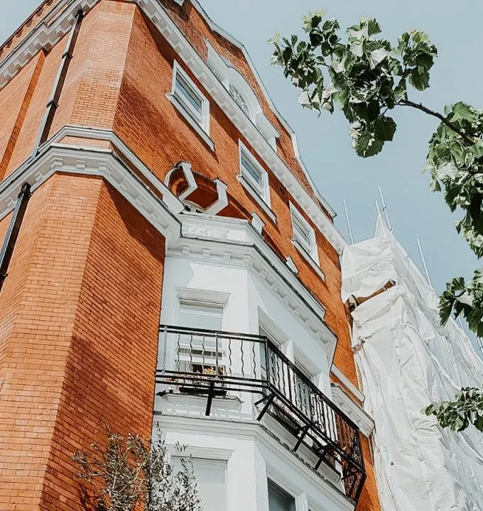Brick building with a decorative balcony, featuring white trim, and scaffolding visible on one side for renovation work.