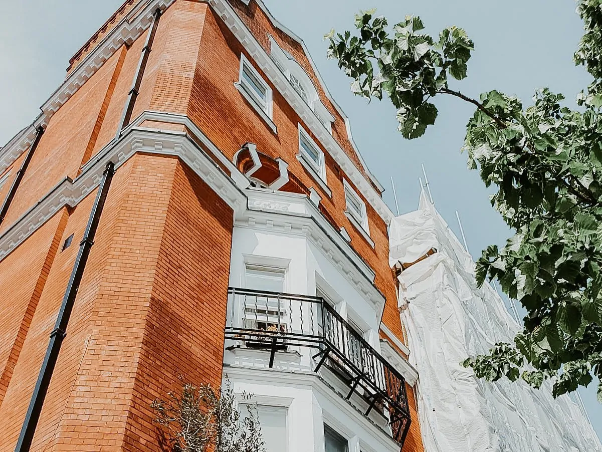 Brick building with a decorative balcony, featuring white trim, and scaffolding visible on one side for renovation work.