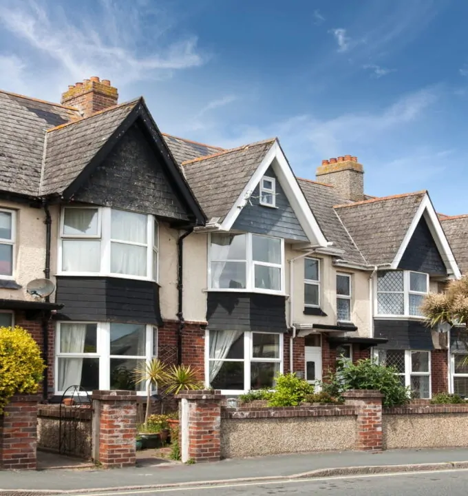 Row of period terraced houses with gabled roofs, bay windows, front gardens, and brick boundary walls along a residential street.